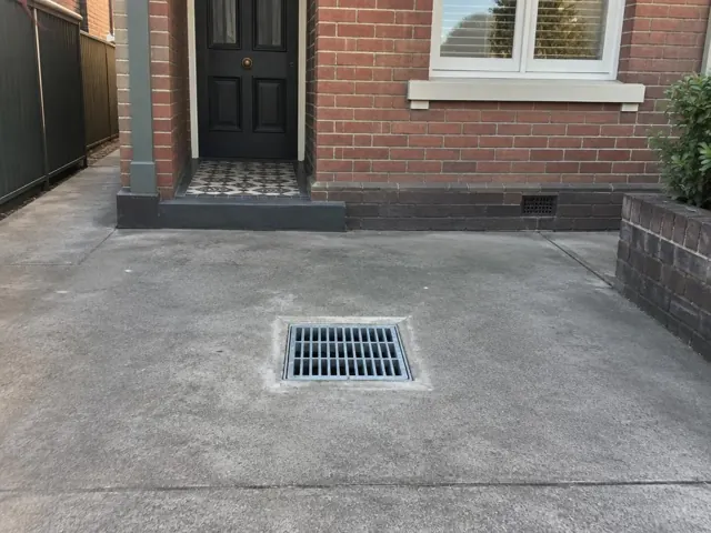 Outdoor drain grate in a concrete driveway beside a brick house in Kingsford