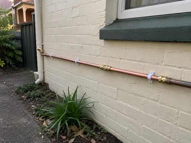 Copper water pipe repaired along a painted brick exterior wall beneath a window in Kingsford
